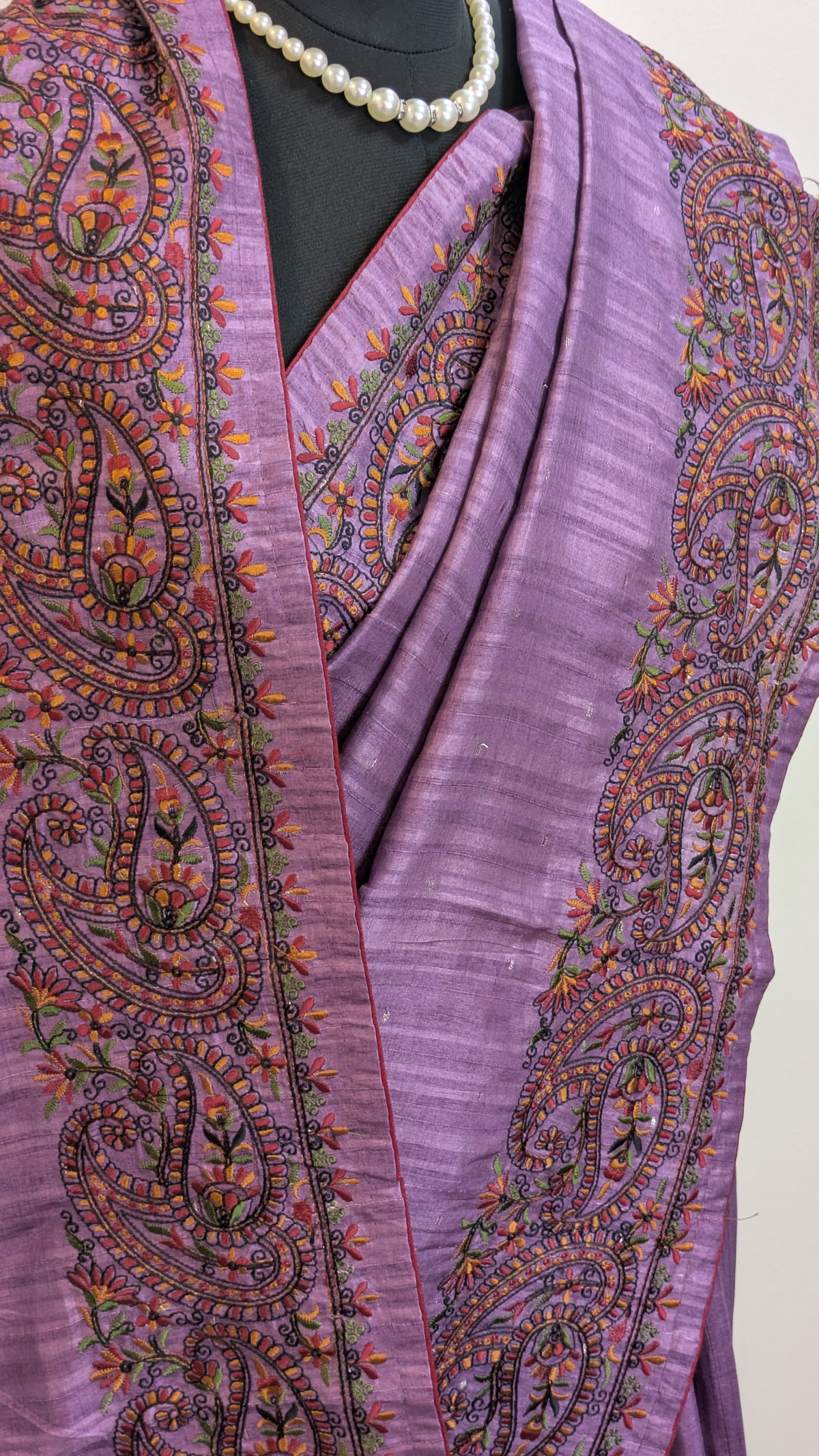 A close-up of a lavender purple embroidered shawl or scarf draped over a black mannequin torso, featuring intricate paisley and floral patterns in orange, green, and black, with a pearl necklace visible at the top.