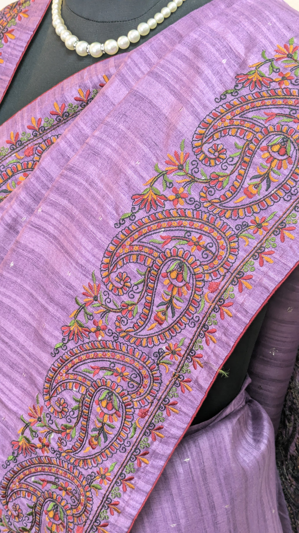 A close-up of a lavender purple saree with detailed multicolor paisley and floral embroidery along the border, styled on a mannequin with a pearl necklace.