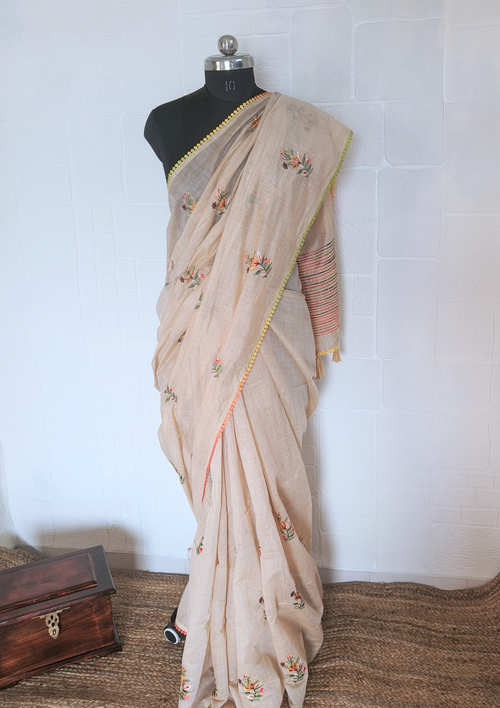 Beige hand-embroidered linen saree with floral motifs and multi-colored lace border draped on a mannequin.